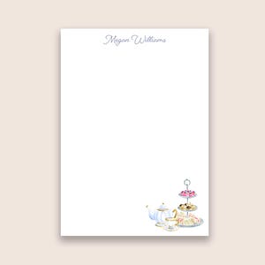 May include: A white rectangular stationery sheet featuring the name Megan Williams in a grey script font at the top center. The bottom right corner displays a watercolor illustration of a white teapot, a teacup, and a three-tiered dessert stand.