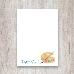 May include: White stationery with a watercolor illustration of a wooden artist's palette, a paintbrush, and the name "Taylor Smith" in teal script. The palette has dabs of yellow, blue, pink, and green paint.