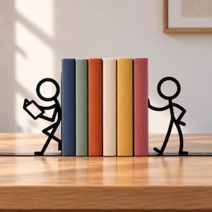 Puede incluir: Un par de sujetalibros de metal negro con siluetas de figuras de palo. Una figura lee un libro abierto, mientras que la otra parece empujar la fila de cinco libros. Los libros tienen cubiertas lisas en azul, verde, naranja, crema y amarillo.