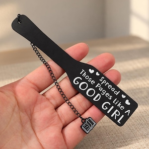 May include: A black metal bookmark featuring a paddle shape with the white text "Spread Those Pages Like A GOOD GIRL" and small heart icons. A thin black chain is attached to the top, ending in a small charm shaped like an open book.