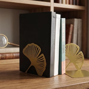 Peut inclure: Une paire de serre-livres en métal doré avec un motif de feuille de ginkgo découpé. Les serre-livres sont placés de chaque côté d'une petite pile de livres, une feuille étant fixée sur le côté d'un livre noir et l'autre posée séparément.