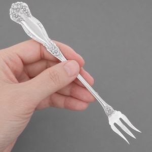 May include: A silver-toned metal cocktail fork featuring an ornate floral embossed pattern on the handle and a smaller floral detail at the base of the three-pronged tines. The utensil has a polished, reflective finish and a slender, tapered stem.