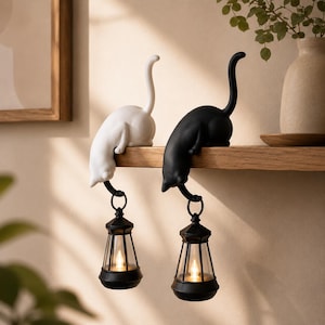 May include: Two cat-shaped ornaments, one white and one black, are perched on a shelf. Each cat has its tail extended upwards and holds a small, black, lantern-style light fixture suspended from its front paws, featuring a warm glowing light.