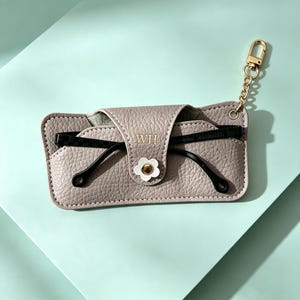 May include: A taupe textured glasses case with a gold-tone metal keychain attachment. The flap features the gold embossed letters WH and a white flower-shaped snap closure. Black glasses are tucked inside the case, resting against the front.