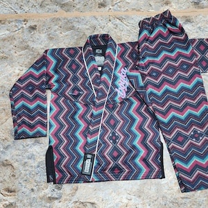 May include: A martial arts gi featuring a dark fabric with a repeating chevron pattern in shades of teal, magenta, and black. The jacket includes a white trim, the text Boyka embroidered in pink on the chest, and a black label with white text.