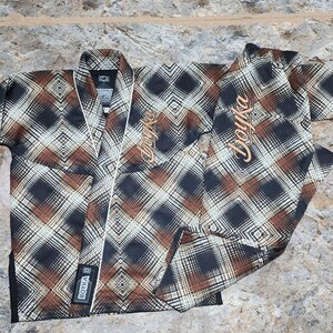 May include: A martial arts gi featuring a black, brown, and white geometric plaid pattern. The jacket has a white trim along the lapel and the text Boyka embroidered in tan on the chest and pant leg. A black Boyka brand patch is on the lower front.