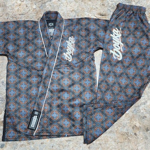 May include: A martial arts gi featuring a dark blue, light blue, and orange geometric pattern. The jacket has a white lapel and a black tag with the text BOYKA. The word Boyka is embroidered in white script on the jacket chest and pant leg.