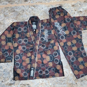 May include: A black jiu-jitsu gi featuring a repeating pattern of copper and grey circles. The jacket has a white trim along the lapel and the text "boyka" in white script on the chest and pants. A black label with "boyka" is on the lower lapel.