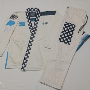 Op de afbeelding: Een witte vechtsport gi-set bestaande uit een jas met een marineblauwe en witte polkadot revers, blauwe en zwarte geborduurde letters op de mouw, en kleine vredes- en gendersymbolen. De bijpassende broek bevat een marineblauwe en witte polkadot patch.