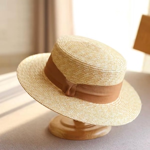 May include: A light tan straw hat with a flat top and a wide brim. The hat features a smooth, solid brown ribbon band wrapped around the base of the crown, tied in a small knot at the front. The woven texture of the straw is visible throughout.