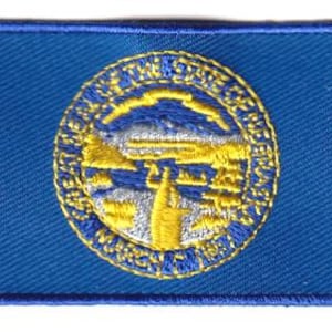 May include: A rectangular embroidered patch featuring the blue flag of Nebraska. The centre displays the state seal in yellow and silver thread, depicting a blacksmith, a steamboat on a river, mountains, and the text Great Seal of the State of Nebraska.