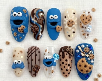 May include: A set of ten press-on nails featuring 3D designs of blue furry characters, chocolate chip cookies, chocolate bars, milk splashes, and lollipops. The nails include various textures like brown chocolate drips, white milk patterns, and blue fur effects with small star and heart accents.