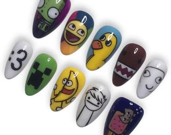 May include: A set of eleven almond-shaped press-on nails featuring various colorful cartoon and internet meme graphics, including a green alien, a yellow duck, a brown creature with sharp teeth, a banana, and a pixelated cat on a dark blue background.