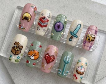 May include: A set of ten press-on nails featuring pixel art designs. The nails display various icons including a gold helmet, TNT block, purple eye, blue sword, bee, treasure chest, fox, red heart, trident, and a birthday cake with flowers.