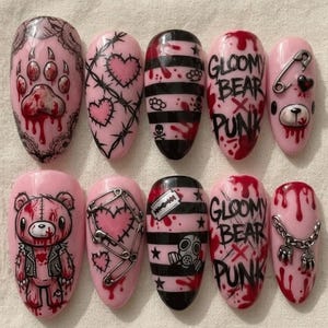 May include: A set of ten almond-shaped press-on nails featuring pink and black designs. The art includes a bloody paw print, barbed wire hearts, black stripes with skulls, the text GLOOMY BEAR PUNK, safety pins, a teddy bear, and a gas mask.
