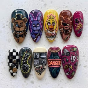 May include: A set of ten press-on nails featuring detailed illustrations of characters and icons from a horror video game. Designs include a brown bear, purple rabbit, yellow chicken, brown fox, pizza, security cameras, a cassette tape, and a sign reading DANGER.