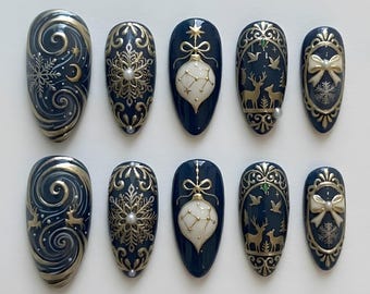 May include: A set of ten almond-shaped press-on nails in deep navy blue with intricate gold and white designs. Patterns include swirling lines, snowflakes, reindeer, bows, and hanging ornaments, accented with small white pearls.