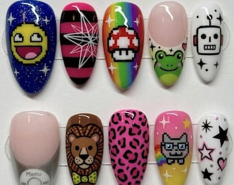 May include: A set of ten press-on nails featuring diverse designs including a yellow pixelated face on blue glitter, pink and black stripes, a rainbow mushroom, a green frog, a white robot, a music player interface, a lion, leopard print, a cat, and stars.