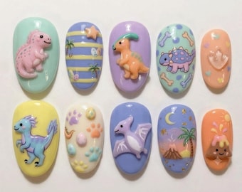 May include: A set of ten press-on nails featuring various dinosaur-themed designs, including 3D figures of dinosaurs, pterodactyls, and volcanoes, alongside patterns of palm trees, stars, moons, and paw prints in pastel shades of blue, pink, and yellow.