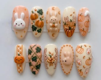 May include: A set of ten almond-shaped press-on nails featuring a fall theme. The designs include 3D white and peach bunnies, orange pumpkins, green leaves, and paw prints on cream, peach, and light pink backgrounds with various star and moon accents.