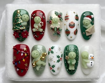 May include: A set of ten press-on nails featuring Christmas themes. Designs include a green glitter nail with a tree, a red nail with a character holding a candy cane, a white nail with holly, a white nail with ornaments, and a green nail with a sleeping character. Other nails feature string lights, a character with gold bells, holly patterns, a character with a tree, and a character with a snowman.