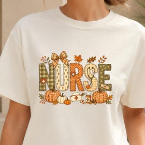 Può includere: Una t-shirt a maniche corte color crema con la parola NURSE in grandi lettere stampate. Il design include motivi autunnali come zucche, foglie, uno stetoscopio, una cuffia da infermiere e un piccolo fiocco nei toni dell'arancione e verde.
