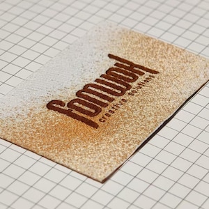 Puede incluir: Un parche rectangular con un fondo degradado de purpurina blanca y dorada, con el texto bordado en marrón oscuro "fomuoh" y el texto más pequeño "creative solutions" colocado en diagonal sobre la superficie.