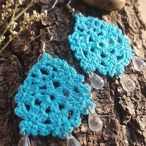 Può includere: Un paio di orecchini all'uncinetto color turchese brillante con un motivo circolare simile al pizzo e bordi smerlati. Ogni orecchino è decorato con tre perline trasparenti a forma di goccia appese a ganci in metallo color argento.