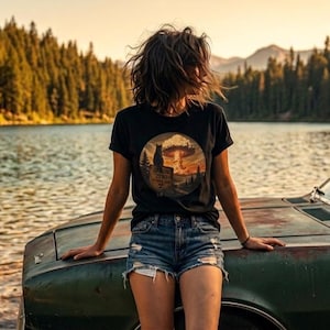 May include: A black short-sleeve t-shirt featuring a circular graphic print on the chest. The design depicts a silhouette of a cat sitting on a rock overlooking a landscape with pine trees and a large mushroom cloud under a sunset sky.
