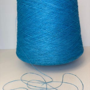 Op de afbeelding: Een grote, conische klos met levendig, felblauw garen. De draad is in een kruislings patroon rond de centrale kern gewikkeld, waarbij een enkele losse streng van hetzelfde blauwe garen vanaf de basis uitsteekt en op het oppervlak eronder krult.