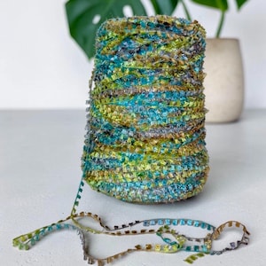 Può includere: Un cono di filato fantasia testurizzato caratterizzato da una tavolozza di colori variegati in verde acqua, verde lime e argento metallizzato. Il filato ha una struttura piatta a nastro con piccoli segmenti di frange distanziati.