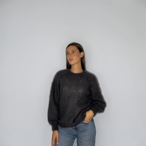 Può includere: Un maglione a maniche lunghe grigio antracite scuro con una finitura in maglia testurizzata e soffice, caratterizzato da un classico girocollo e una vestibilità rilassata, abbinato a jeans blu con chiusura a bottoni e zip.