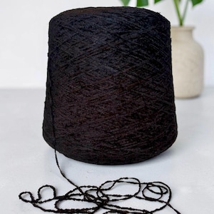 Pode incluir: Um grande cone cilíndrico de fio preto escuro com uma superfície texturizada e ligeiramente irregular. Um único fio do novelo preto estende-se da base do cone e está enrolado de forma solta num padrão emaranhado à frente do cone.