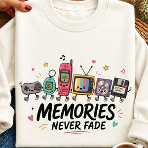 Puede incluir: Una sudadera color crema con un gráfico de tecnología de los años 90 antropomórfica, incluyendo un mando de juego, mascota digital, teléfono móvil, televisión, CD y disquete, con el texto "MEMORIES NEVER FADE" impreso en negro debajo.