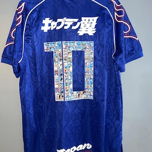 Könnte beinhalten: Ein blaues Kurzarmtrikot mit weißem Kragen und rotem Rand, einem tonalen Flammenmuster und dem japanischen Text キャプテン翼 über einer großen Nummer 10, die mit Anime-Charaktergrafiken gefüllt ist. Das Wort Japan ist unten in Weiß aufgedruckt.