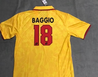 May include: A yellow short-sleeved jersey featuring a subtle geometric pattern. The back displays the name BAGGIO in black lettering above the number 18 in red with black outlines. The collar and sleeve cuffs are trimmed in red and dark yellow.