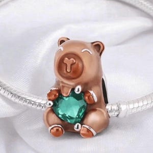 May include: A rose gold-toned bear charm bead featuring a smiling face and holding a faceted, heart-shaped emerald green gemstone. The charm is threaded onto a silver-toned snake chain bracelet, showcasing a polished metallic finish throughout.