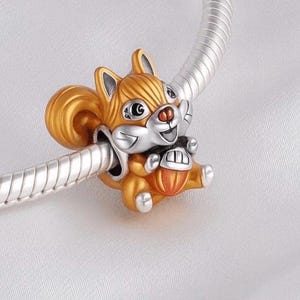 May include: A silver charm bead shaped like a squirrel with orange enamel detailing, holding a small acorn. The squirrel has large eyes, a bushy tail, and is threaded onto a silver snake chain bracelet, showcasing a polished metallic finish.