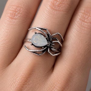 May include: A silver-toned metal ring featuring a spider design with a pear-shaped, iridescent white stone set in the centre of the spider's body. The spider has eight articulated legs that extend along the band of the ring.