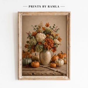 以下が含まれることがあります： PRINTS BY RAMLAと題された額入りアートプリント。ベージュの陶器製ピッチャーに活けられた白とオレンジの紫陽花、秋の葉、ユーカリのフラワーアレンジメントと、その周りに配置されたオレンジやセージグリーンのカボチャが描かれています。