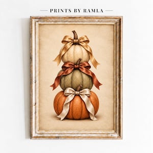 May include: A framed wall art print titled PRINTS BY RAMLA featuring a vertical stack of three pumpkins in cream, sage green, and orange. Each pumpkin is adorned with a satin ribbon bow in matching tones, set against a textured beige background.