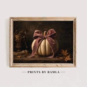 May include: A framed wall art print featuring a light-colored pumpkin tied with a large, dusty rose velvet ribbon. The pumpkin sits on a dark wooden surface alongside scattered dried autumn leaves and a small vase of dried flowers in the background.