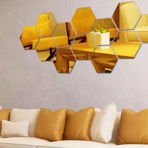 Peut inclure: Un ensemble de sept miroirs muraux hexagonaux disposés en grappe. Chaque miroir individuel présente une surface réfléchissante dorée qui affiche une vue déformée d'une pièce avec des meubles jaunes, un cache-pot carré blanc et une plante verte.