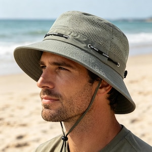 Op de afbeelding: Een olijfgroene bucket hat met een brede rand, een mesh kroongedeelte voor ventilatie en een verstelbare zwarte kinband. De hoed is voorzien van een decoratief zwart koord rond de basis van de kroon met zilverkleurige hardware.