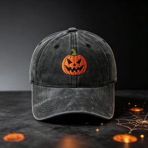 May include: A charcoal grey baseball cap with a distressed, washed-out finish. Centered on the front panel is an embroidered orange jack-o'-lantern pumpkin with a green stem, featuring a jagged, triangular-eyed face in black thread.