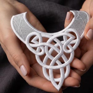 Puede incluir: Una aplicación de cuello bordada en color blanco que presenta un patrón denso e intrincado de nudos celtas. El diseño consiste en bucles y líneas interconectadas con un acabado de puntada en relieve y texturizado, formando una pieza decorativa.