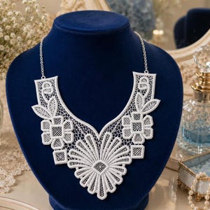 May include: A white lace statement necklace featuring an intricate floral and geometric pattern. The collar-style piece is constructed from delicate white thread work, showcasing a large central sunburst motif flanked by smaller square and leaf designs.