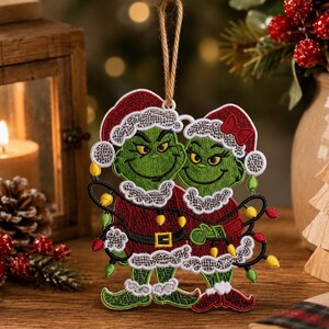 May include: A hanging embroidered ornament featuring two green characters wearing red Santa hats and matching outfits with white lace-like trim. They are wrapped in a string of colorful Christmas lights with yellow, red, and green bulbs.