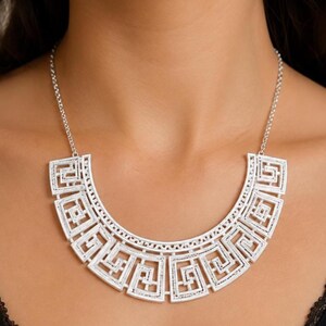 May include: A silver-toned bib necklace featuring a prominent semi-circular pendant with a repeating geometric Greek key pattern. The necklace is suspended from a delicate silver chain and showcases a textured, matte metallic finish throughout.