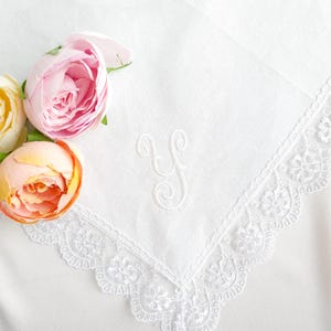 May include: A white square handkerchief featuring a delicate white lace border with a floral pattern and a cursive letter Y embroidered in the center. Three artificial flowers in shades of pale yellow, soft pink, and peach are placed on the left.